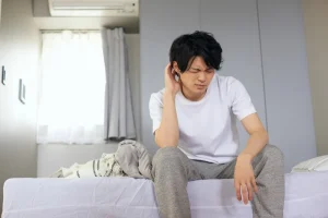man sits at edge of bed, touching ear
