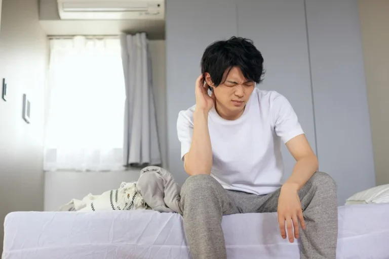 man sits at edge of bed, touching ear