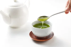 spoonful of green tea poured into white mug on wooden saucer beside white teapot